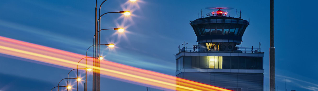 Airport control tower at night with glowing strips of light from passing vehicles, symbolizing time synchronization.